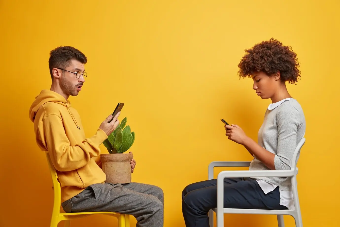 An addicted multiracial couple with internet addiction sit on chairs opposite each other, using their mobile phones for online communication and spending their free time at home.